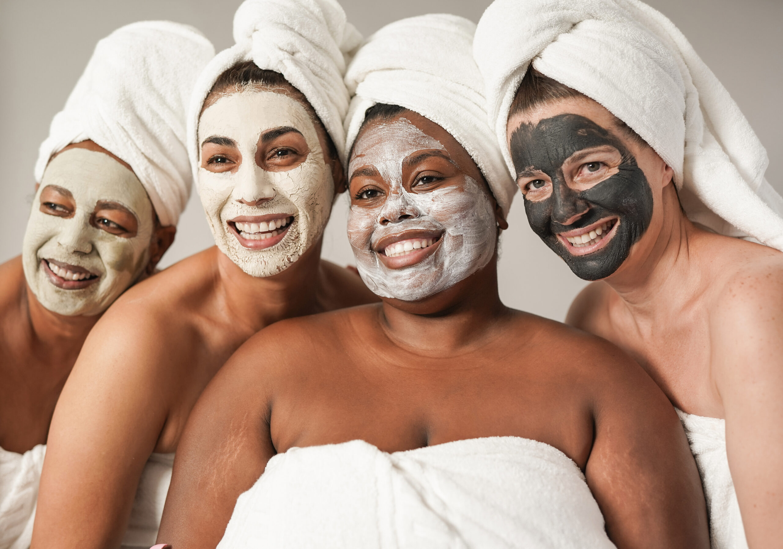 Multiracial women enjoy spa day together with beauty face mask while wearing towels - Multi generational friends with different skin color and bodys