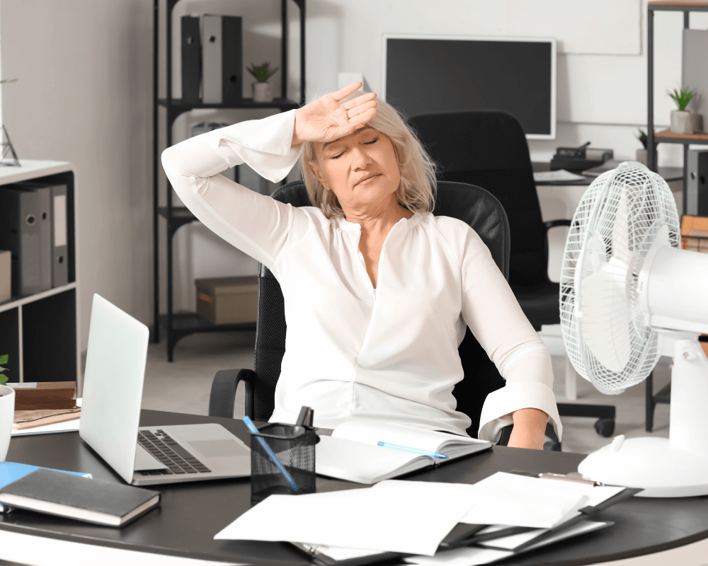 A person seated in an office chair leans back with one hand resting on their forehead, possibly feeling fatigued or hot. A white fan is positioned on the desk, directing air toward the person. The desk features a laptop, an open notebook, a pen holder, and scattered papers. Shelves with binders, potted plants, and another computer monitor are visible in the background, depicting a busy office environment.