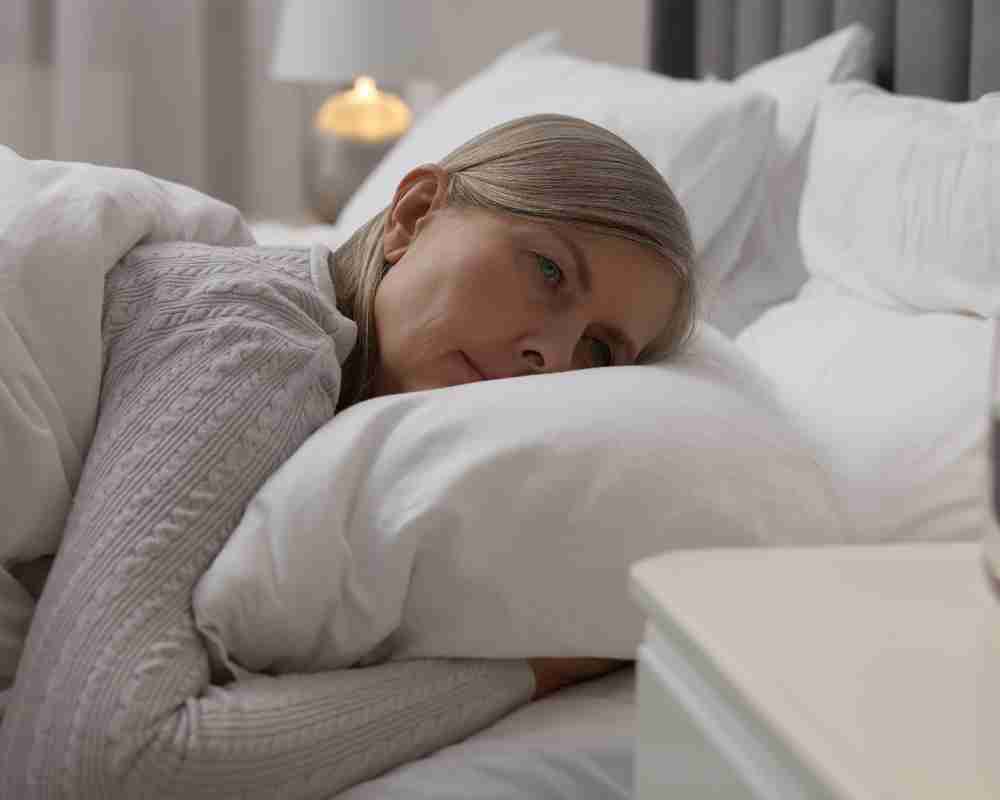 A person lying in bed, resting on a pillow with their face blurred for privacy. They are covered by a white blanket and wearing a light-colored, textured sweater. The background features a bedside table with a lamp emitting warm light, creating a cozy and softly lit atmosphere.